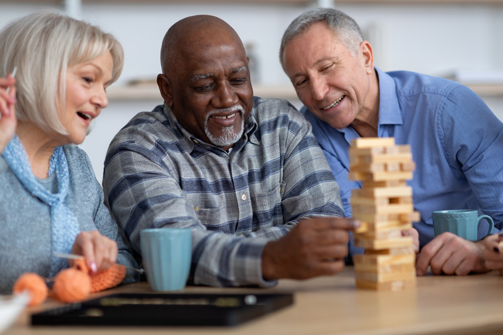 Three older adults playing a puzzle game called Jenga at a table and drinking tea. 