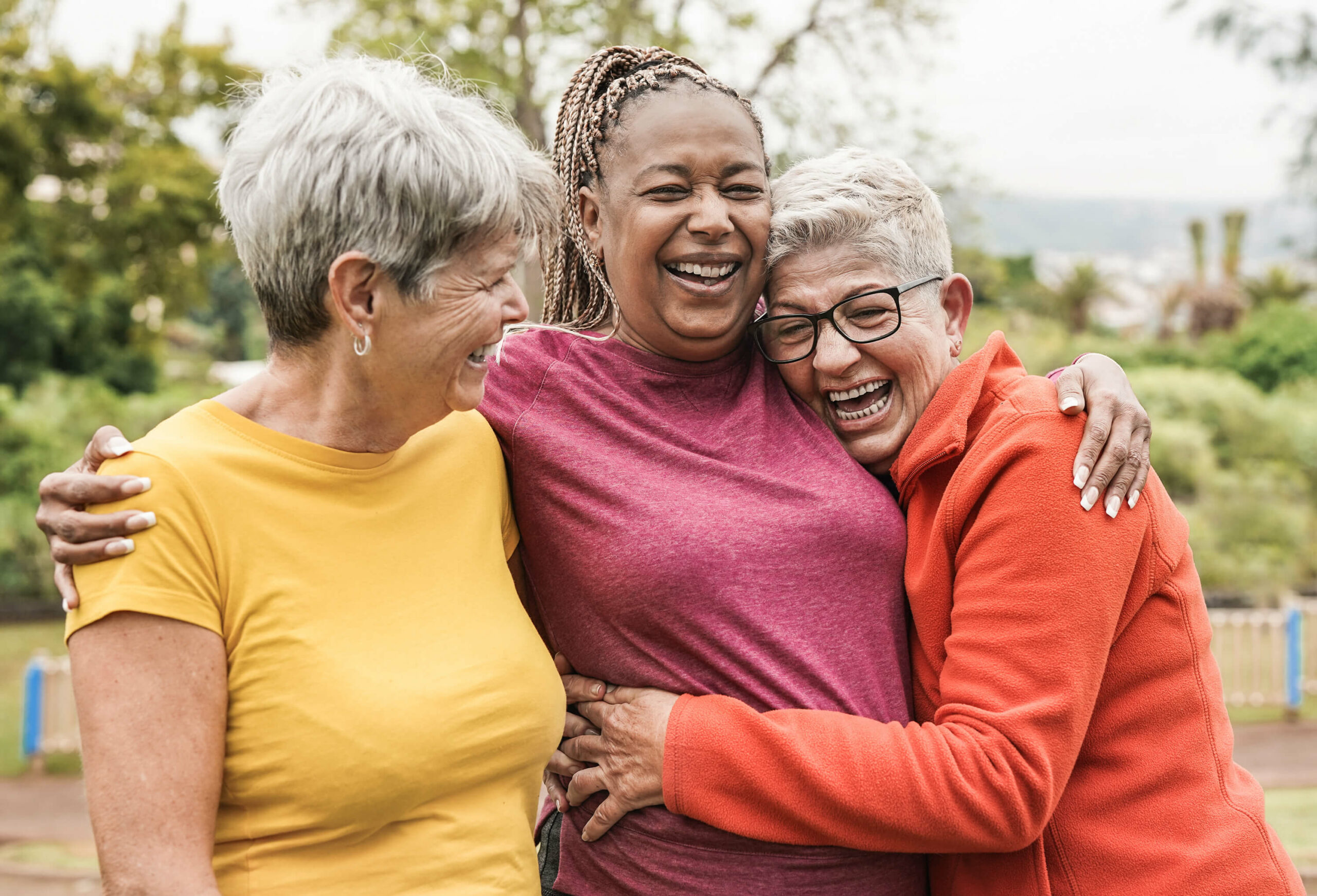 Three older adults women friends outdoors in a park embracing each other and laughing.
