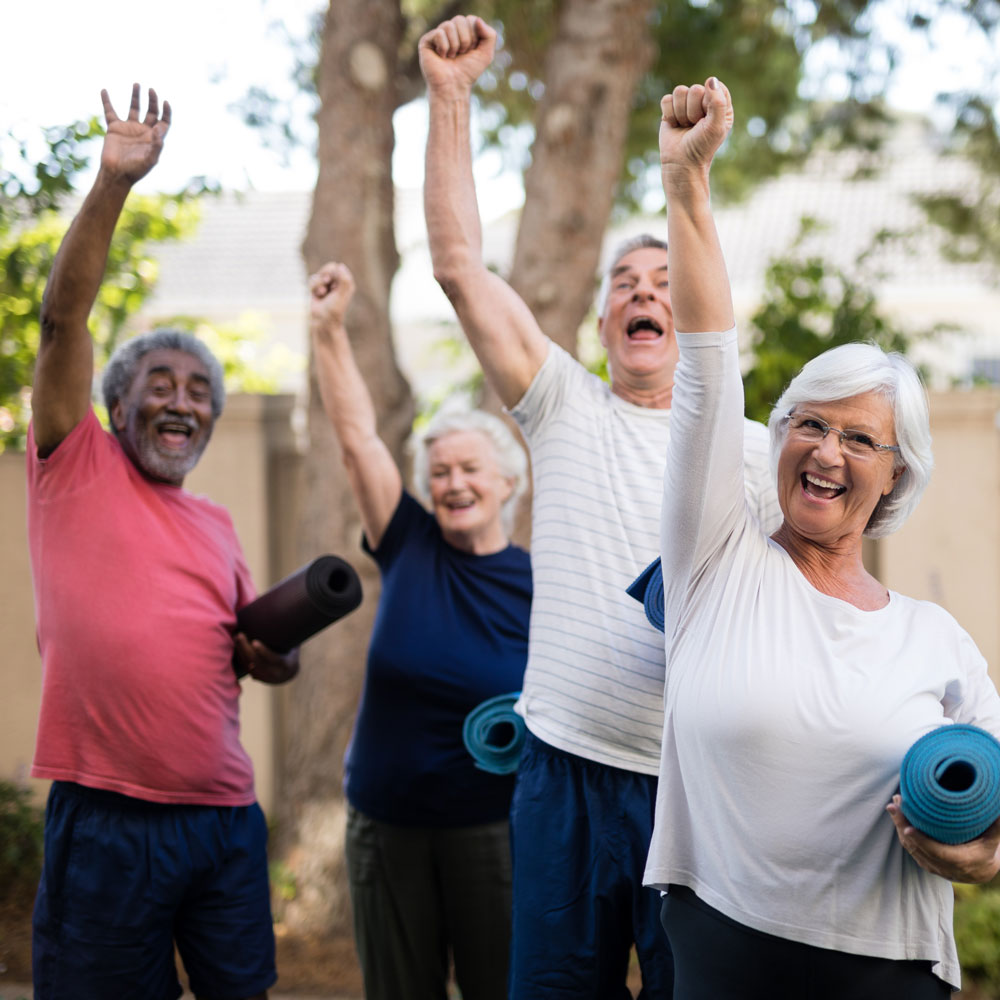 A group of smiling older adults each with one arm enthusiastically raised in celebration and the other arm holding a yoga matt.