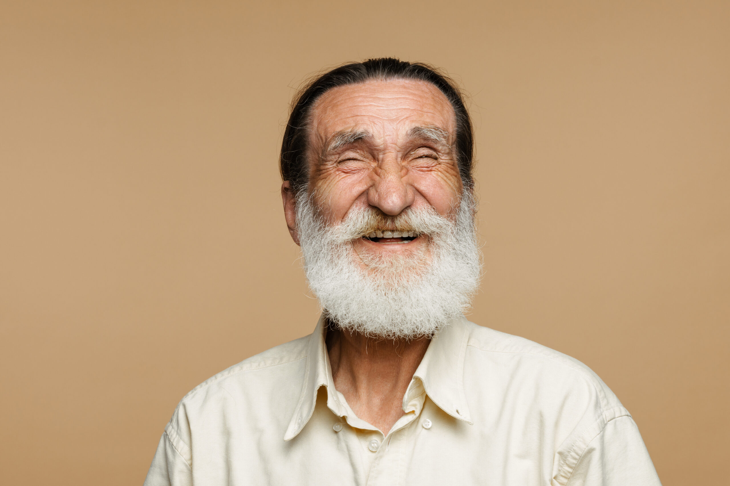 European senior man with beard posing and laughing at camera isolated over beige wall