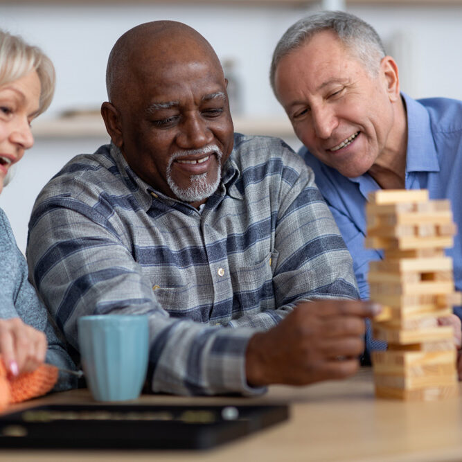 Three older adults playing a puzzle game called Jenga at a table and drinking tea.