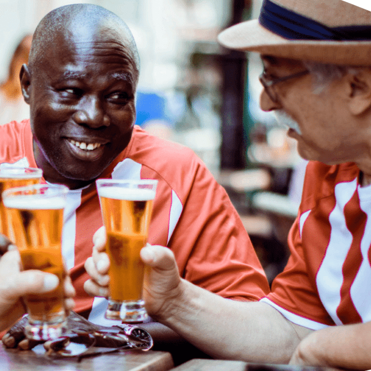 Am man with dark skin, and a man white light skin wearing a hat are holding up beer to cheers.